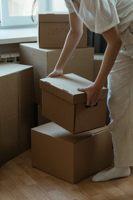 A person wearing light-colored clothing is seen stacking and organizing cardboard boxes in an indoor space, likely part of a house removal process. The boxes vary in size and are made of brown corruga