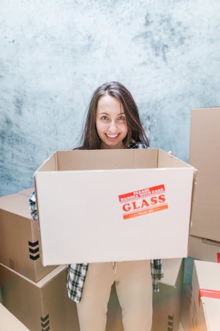 A person wearing light-colored clothing is seen stacking and organizing cardboard boxes in an indoor space, likely part of a house removal process. The boxes vary in size and are made of brown corruga